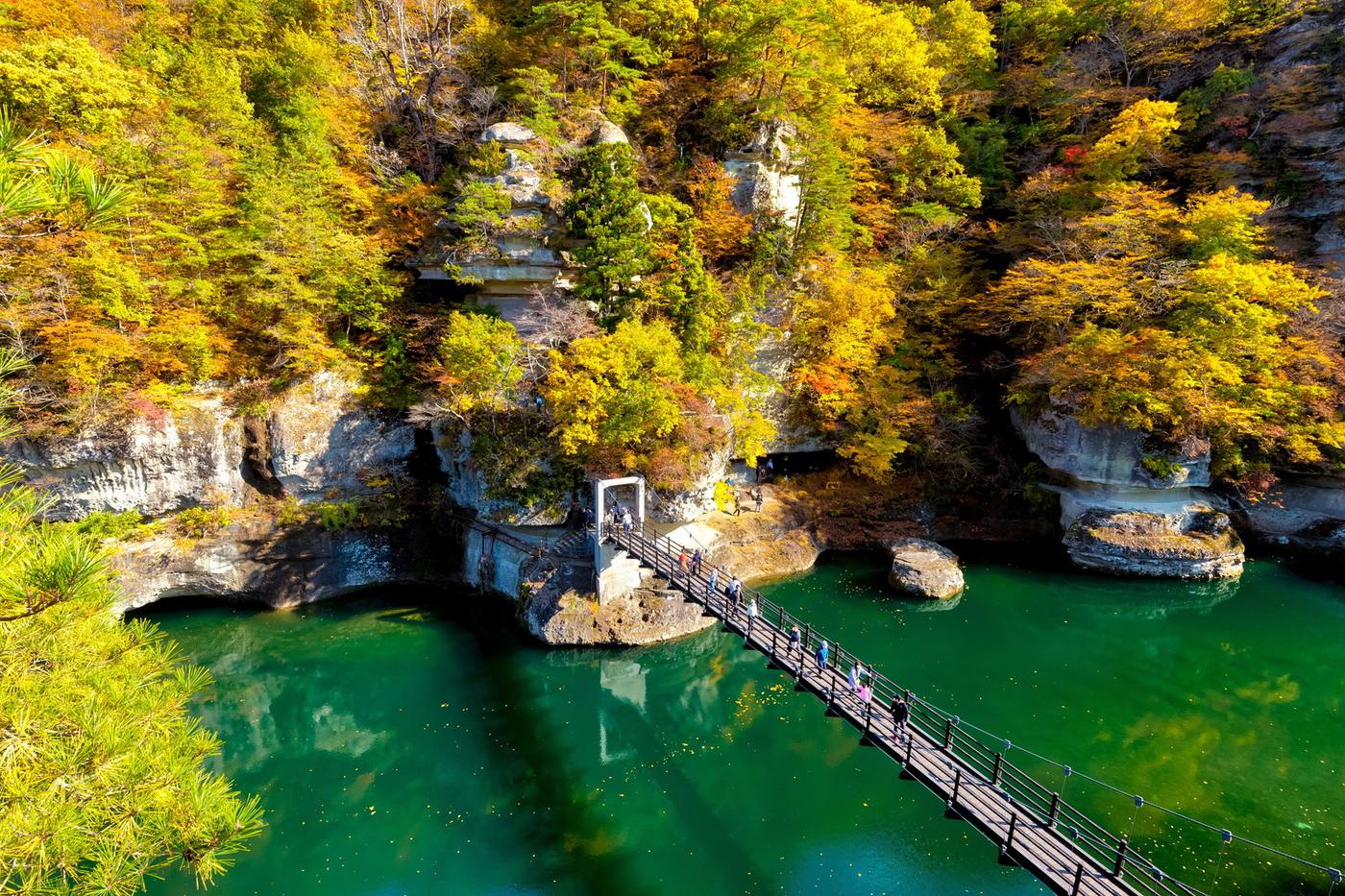 Pale tower-like cliffs and autumn trees above the green Okawa River at To-no-hetsuri, with a suspension bridge crossing the water